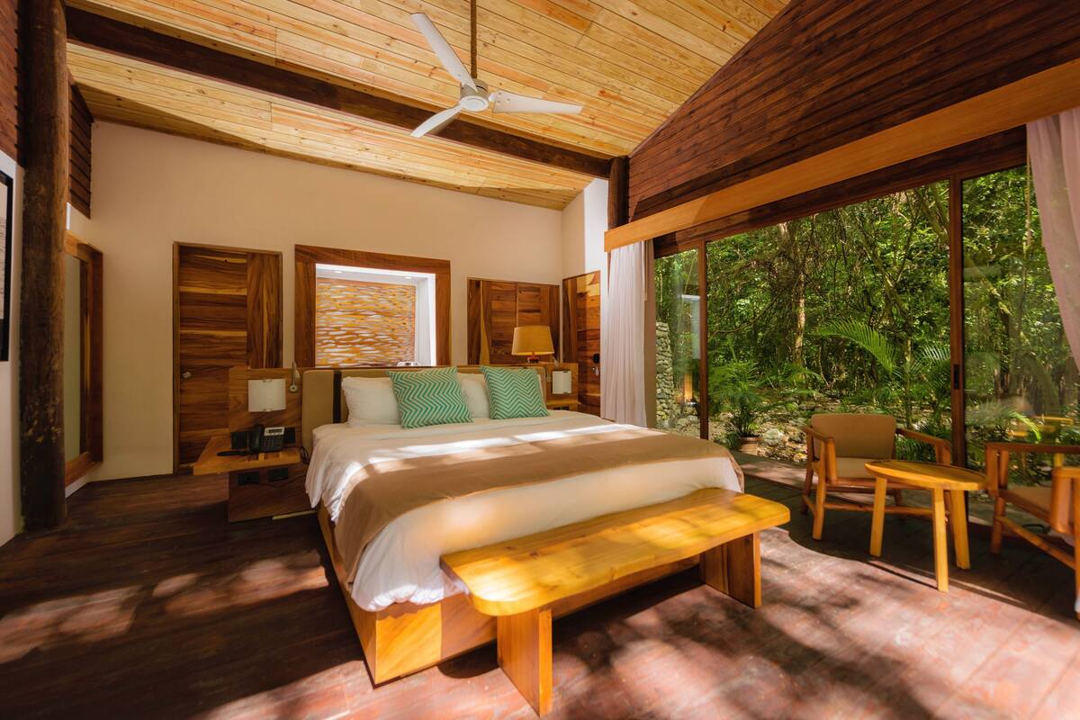 Un dormitorio con cama, mesitas de noche, un banco y vistas al exterior.
