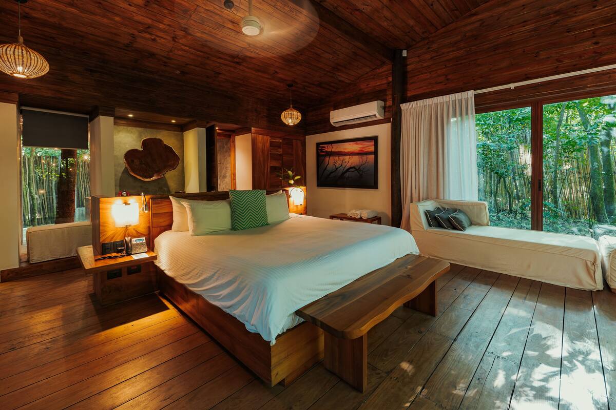 Un dormitorio con suelo de madera, cama, mesitas de noche, un banco, un sofá y un gran ventanal con vistas a árboles.