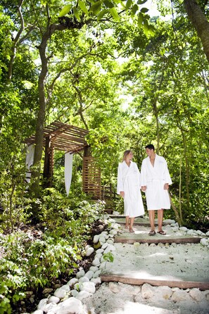 A couple in white robes walking on a stone pathway surrounded by lush greenery and a wooden pergola structure.