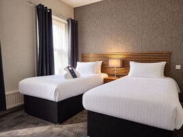 A hotel room with two beds, a wooden headboard, a nightstand with a lamp, and a window with curtains.