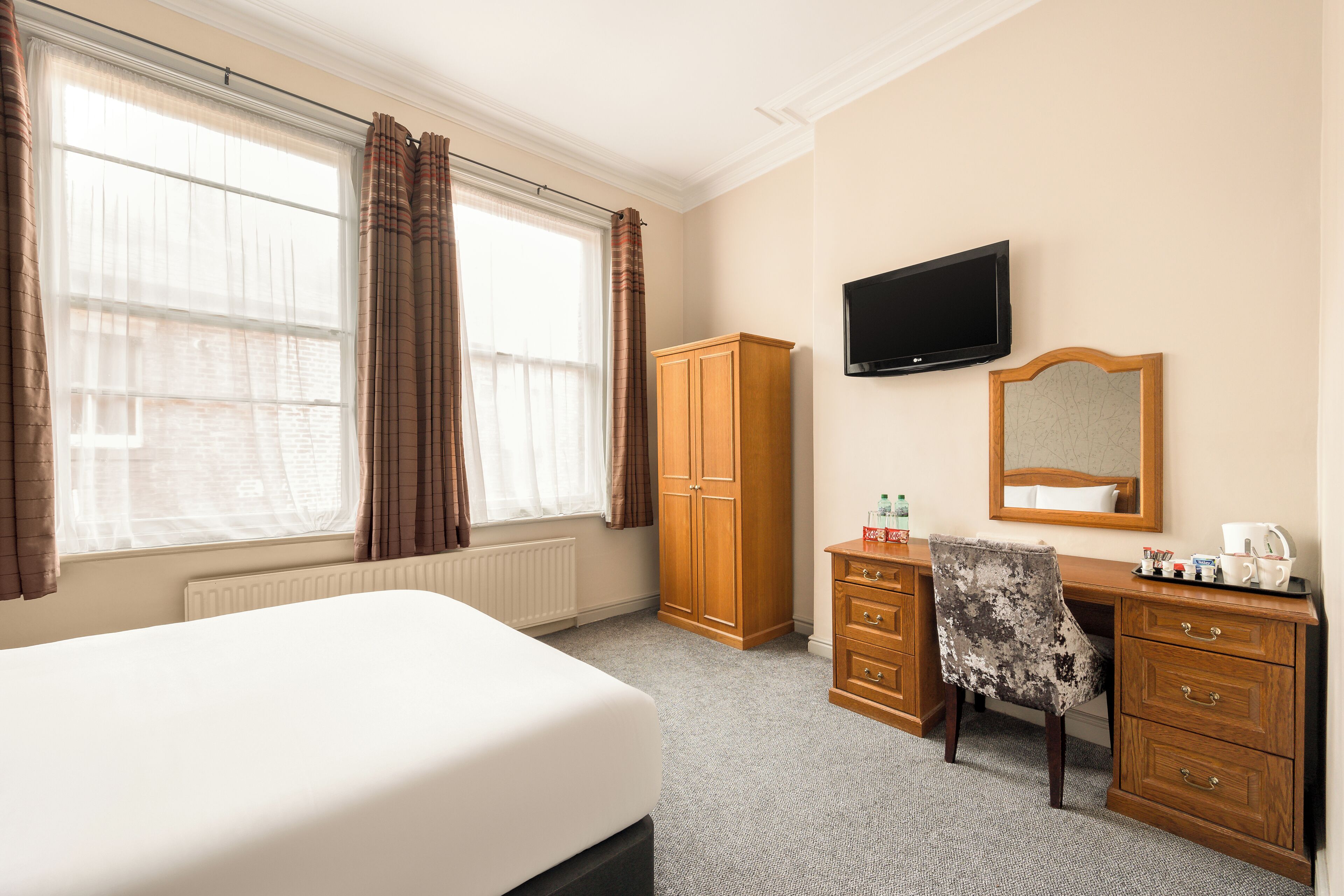 Een hotelkamer met een bed, een houten garderobe, een bureau met stoel, een flatscreen televisie en een groot raam met gordijnen.