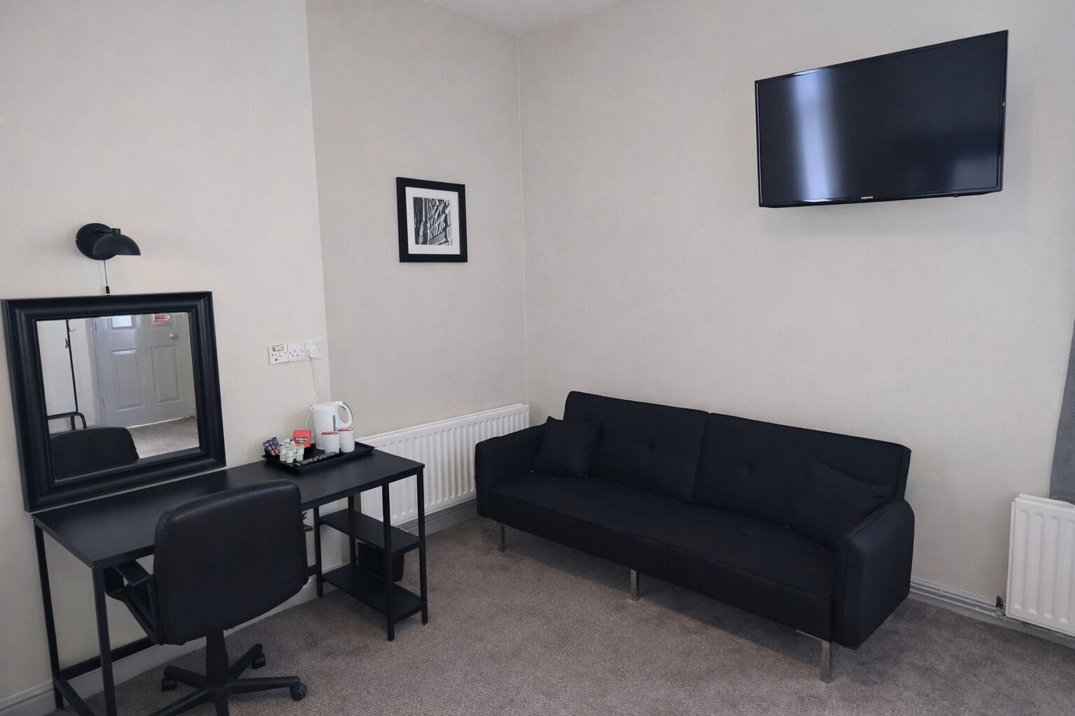 Ein kleiner, moderner Raum mit einem schwarzen Schreibtisch, einem schwarzen Bürostuhl, einer schwarzen Couch und einem festmontierten Flachbildfernseher. An der Wand hängt ein gerahmtes Bild.
