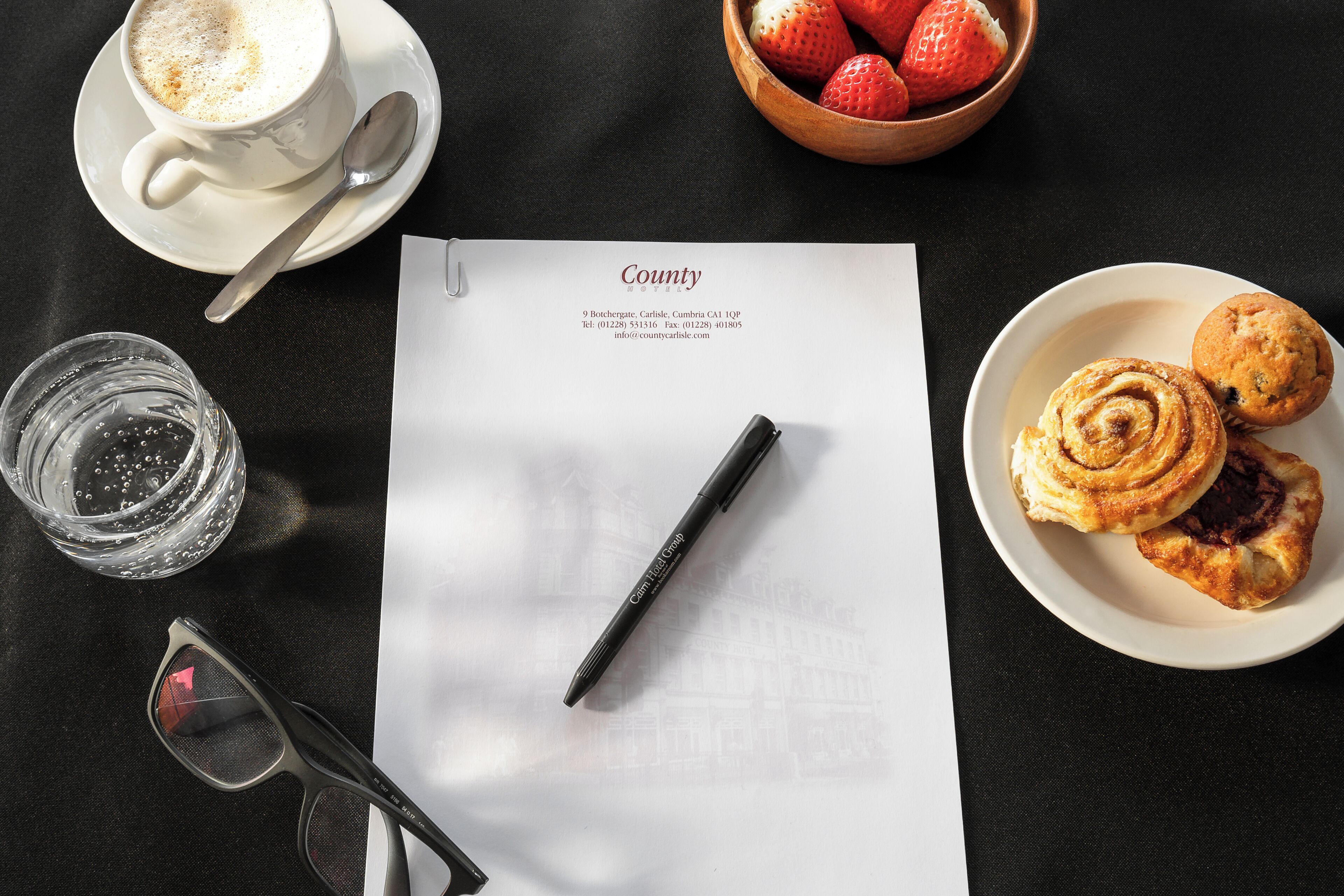 A table with a cup of coffee, a bowl of strawberries, a plate of pastries, a glass of water, a pair of glasses, and a document with a pen.
