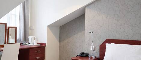 A hotel room with a large bed, a desk with a chair, a small bedside table with a lamp, and a wall with a floral pattern.
