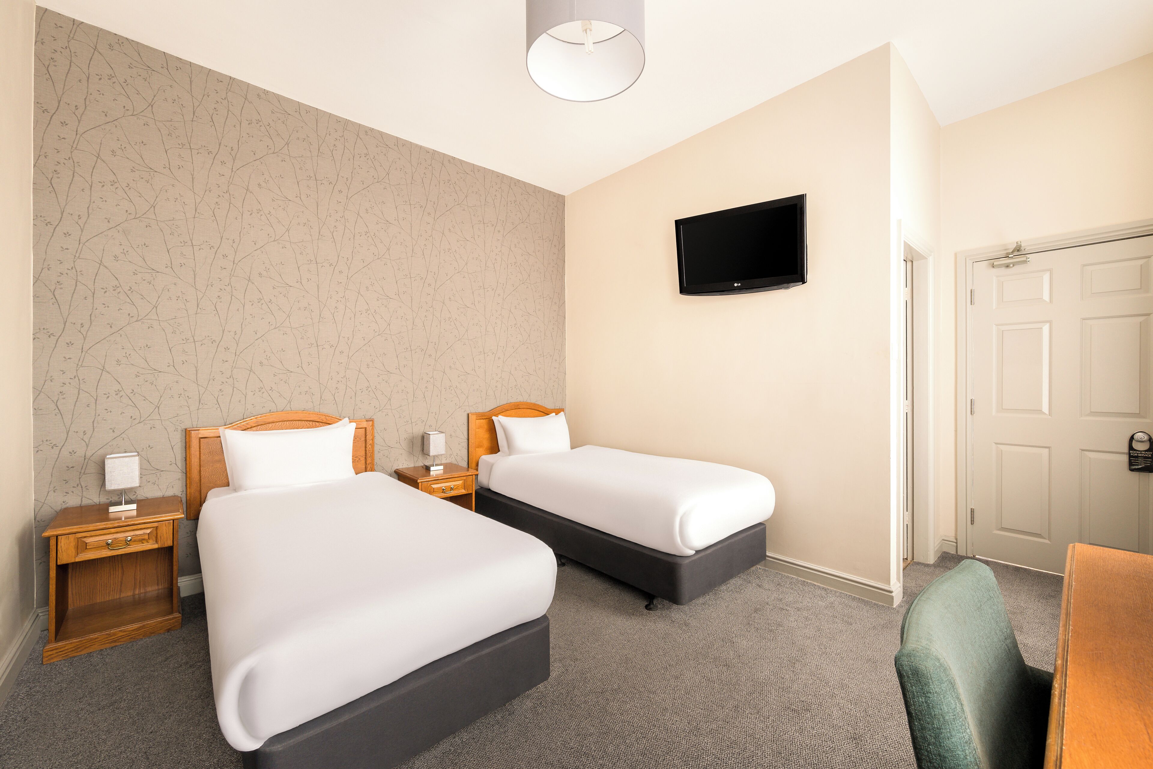 A hotel room with two beds, a TV mounted on the wall, a wooden nightstand with a lamp, and a door.