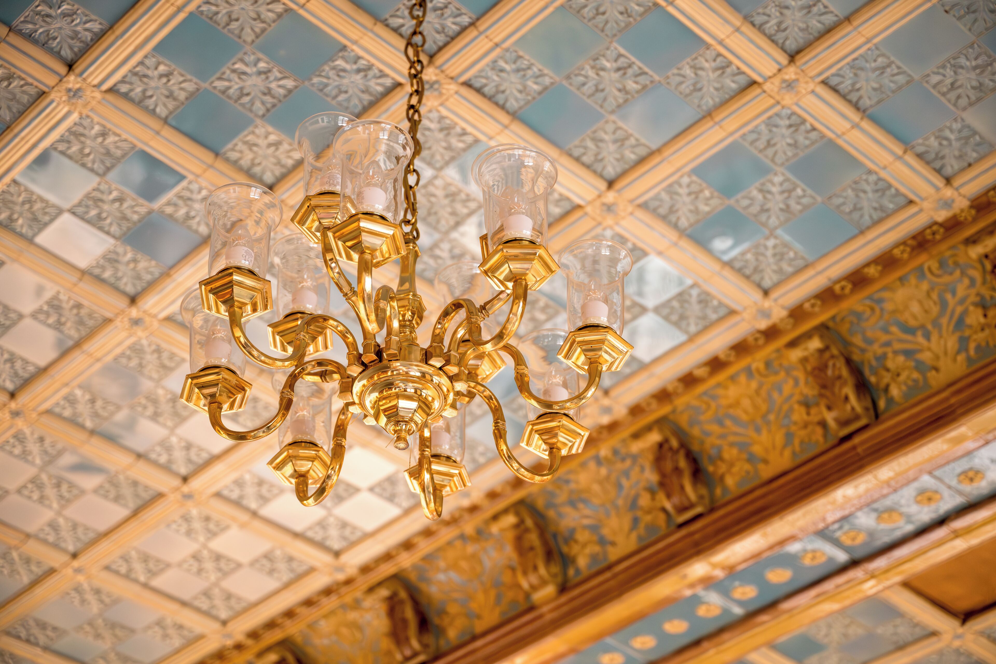Un lustre à plusieurs bras, orné d’abat-jours en verre transparent, suspendu à un plafond décoré de carreaux à motifs.