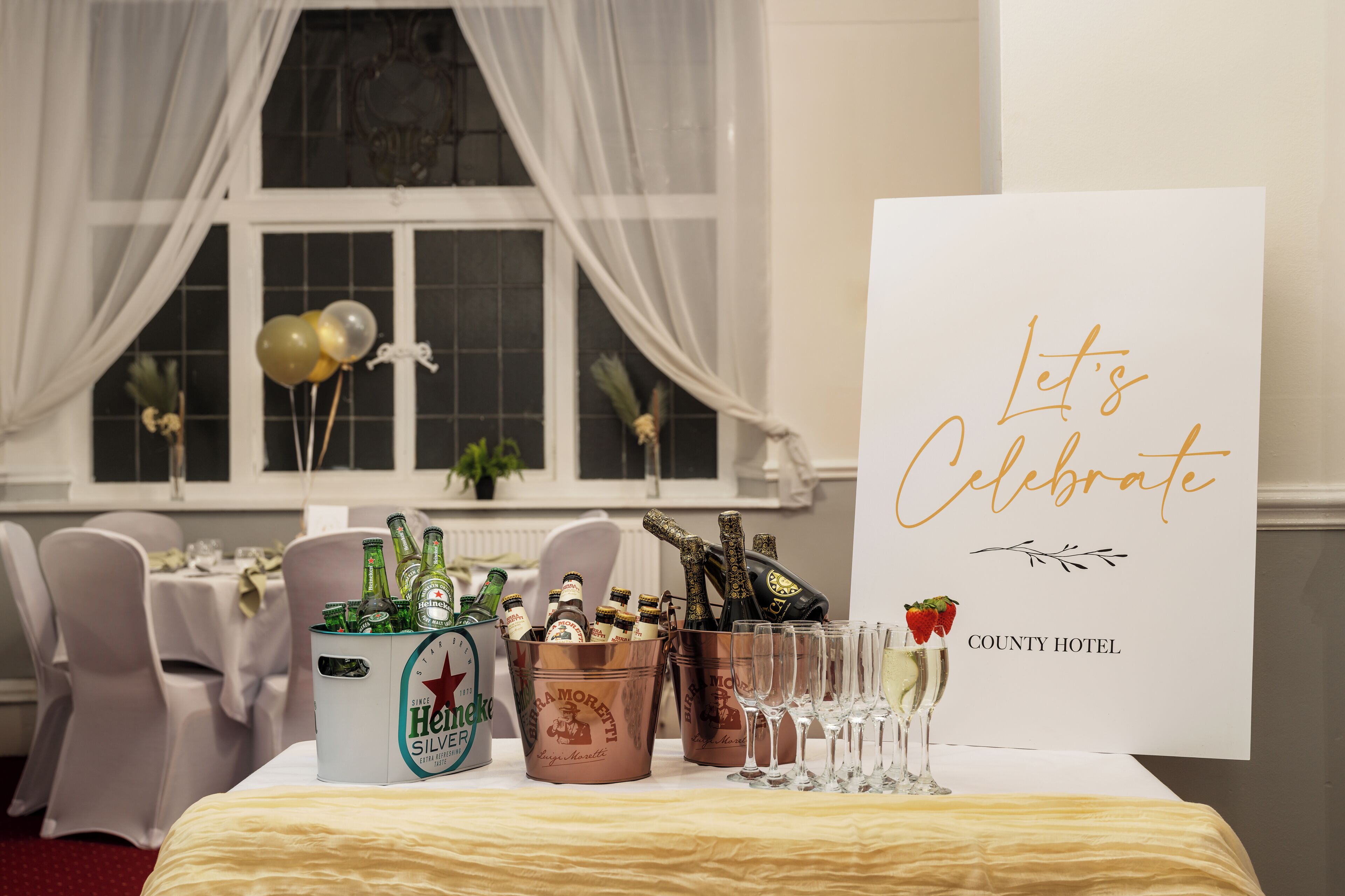 A table set with champagne bottles, glasses, and a sign that reads 'Let's Celebrate' at the County Hotel.
