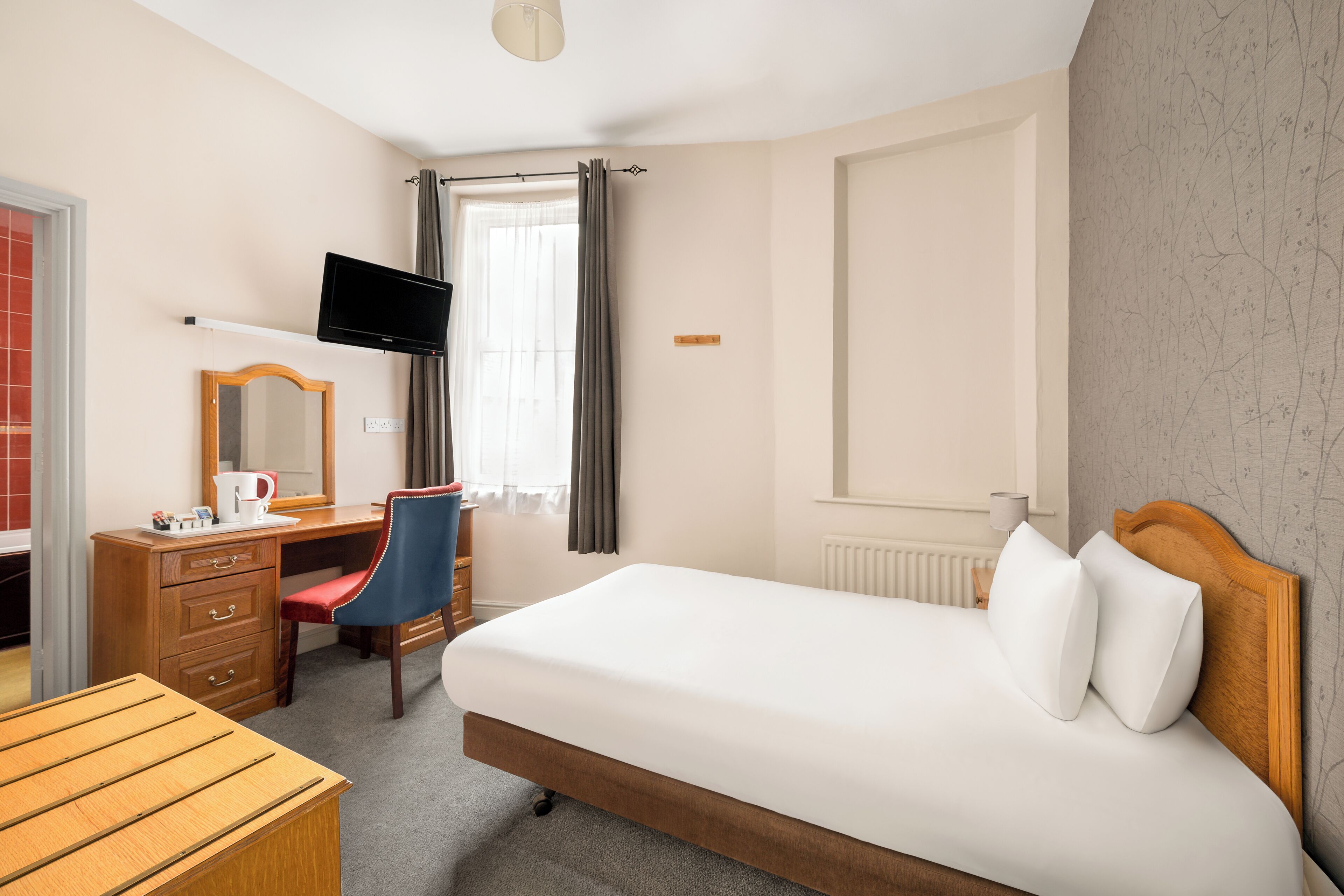 Ein Hotelzimmer mit einem Bett, einem Schreibtisch mit Stuhl, einem Fernseher und einem Fenster mit Vorhängen.