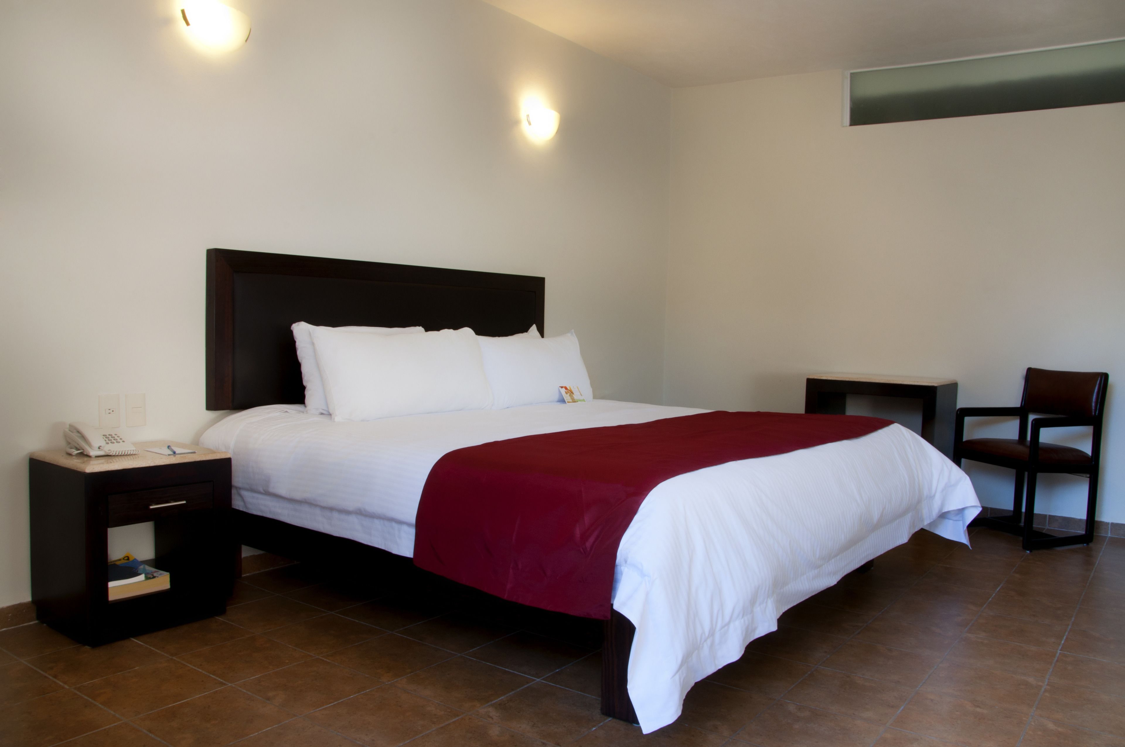 A neatly made bed with white linens and a red blanket, a bedside table with a phone and notepad, and a chair to the right.