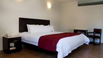 A neatly made bed with white linens and a red blanket, a bedside table with a phone and notepad, and a chair to the right.