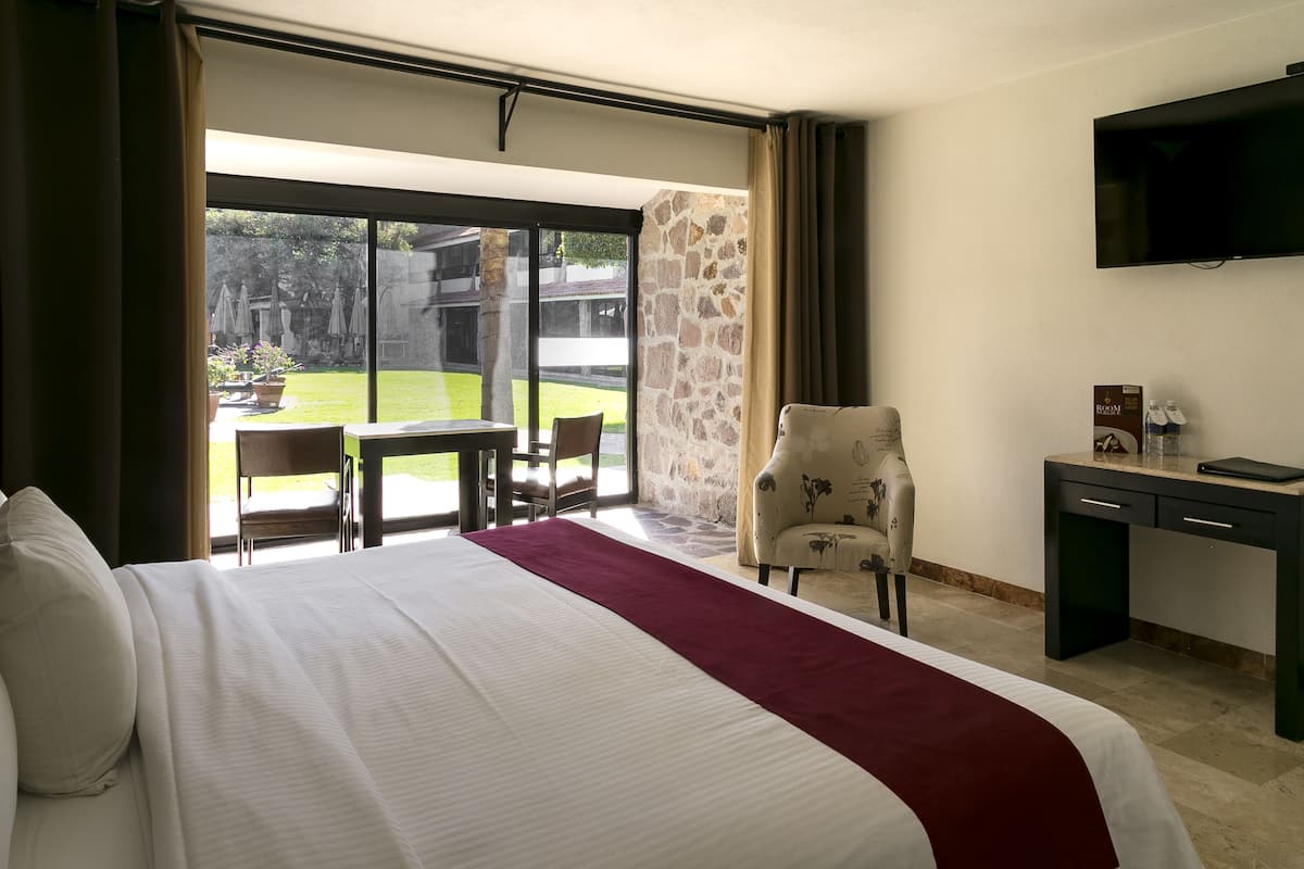 A hotel room with a large bed, a desk, a chair, a TV, and a view of the outdoor area.
