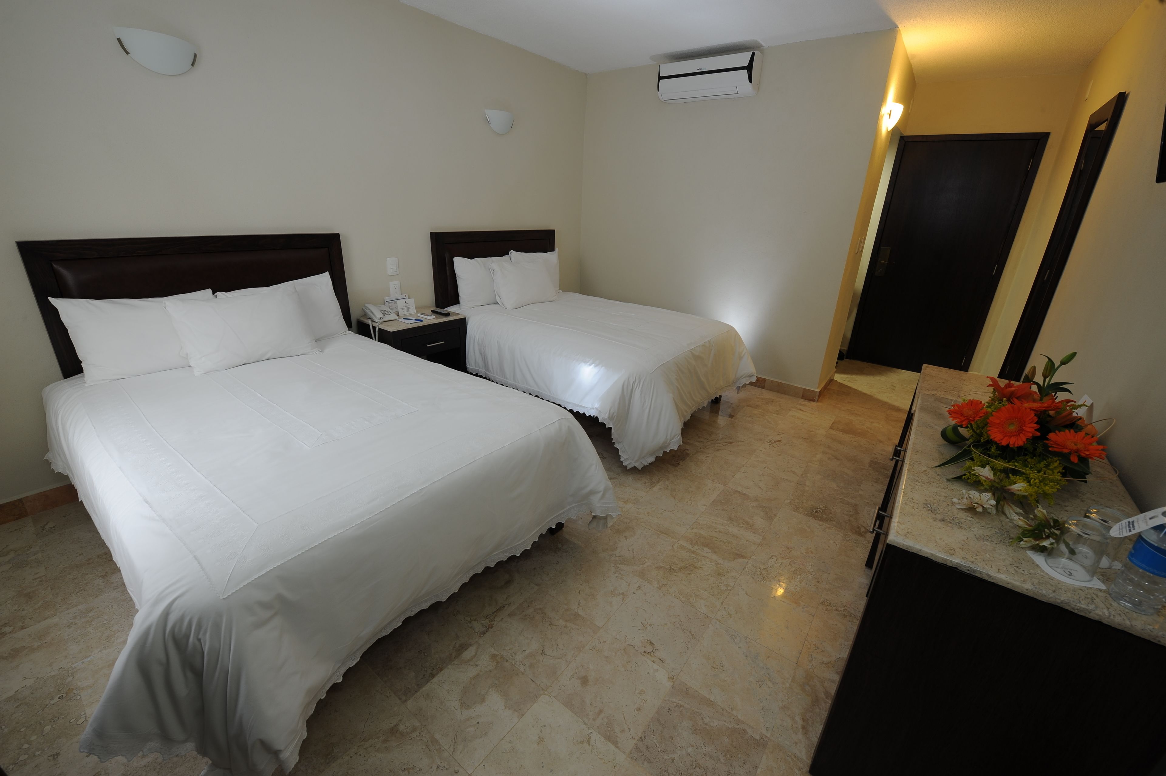 Habitación de hotel con dos camas, una mesita de noche, un escritorio con un jarrón de flores y un equipo de aire acondicionado.