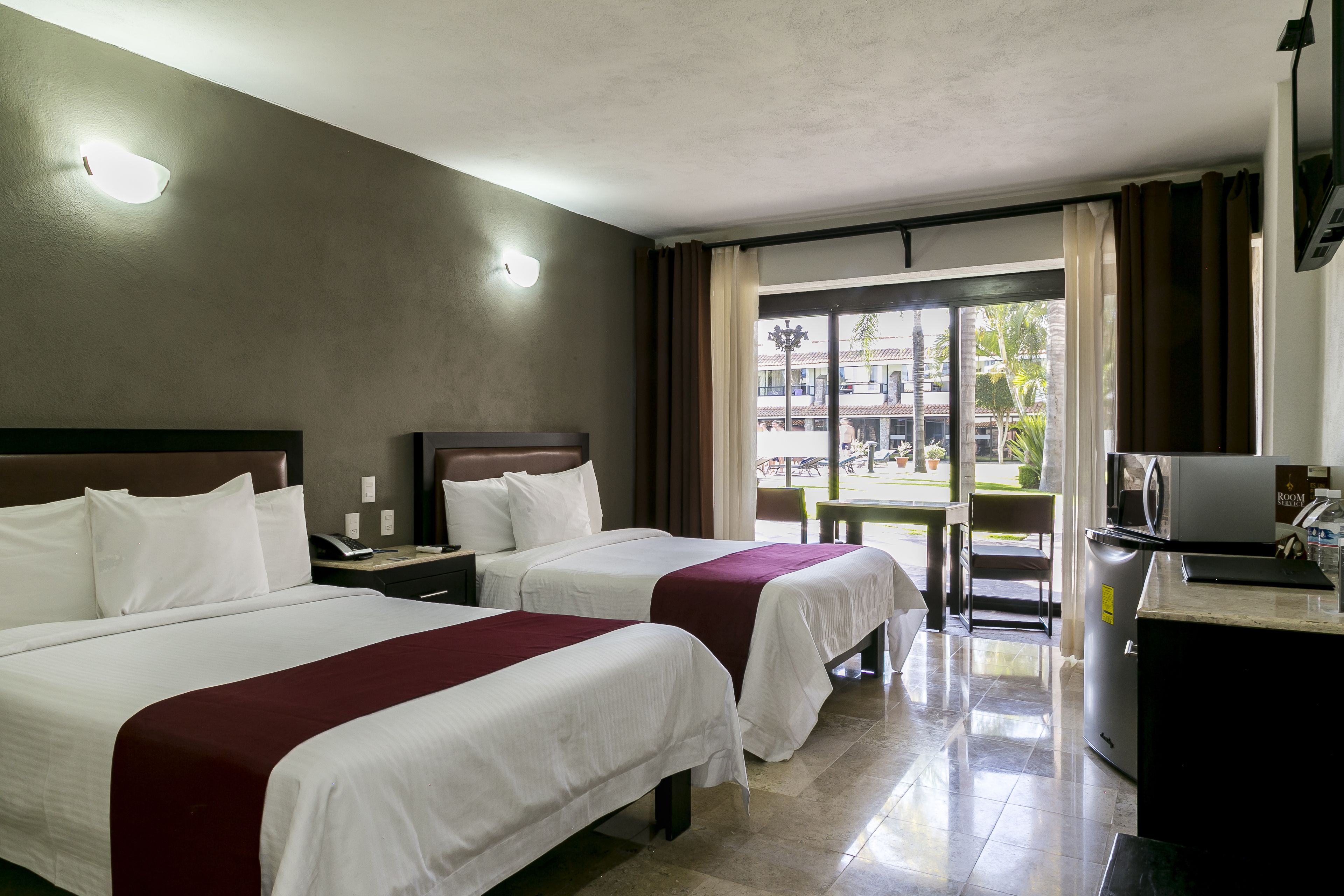 A hotel room with two beds, a desk, a chair, a small table, and a view of the outside.