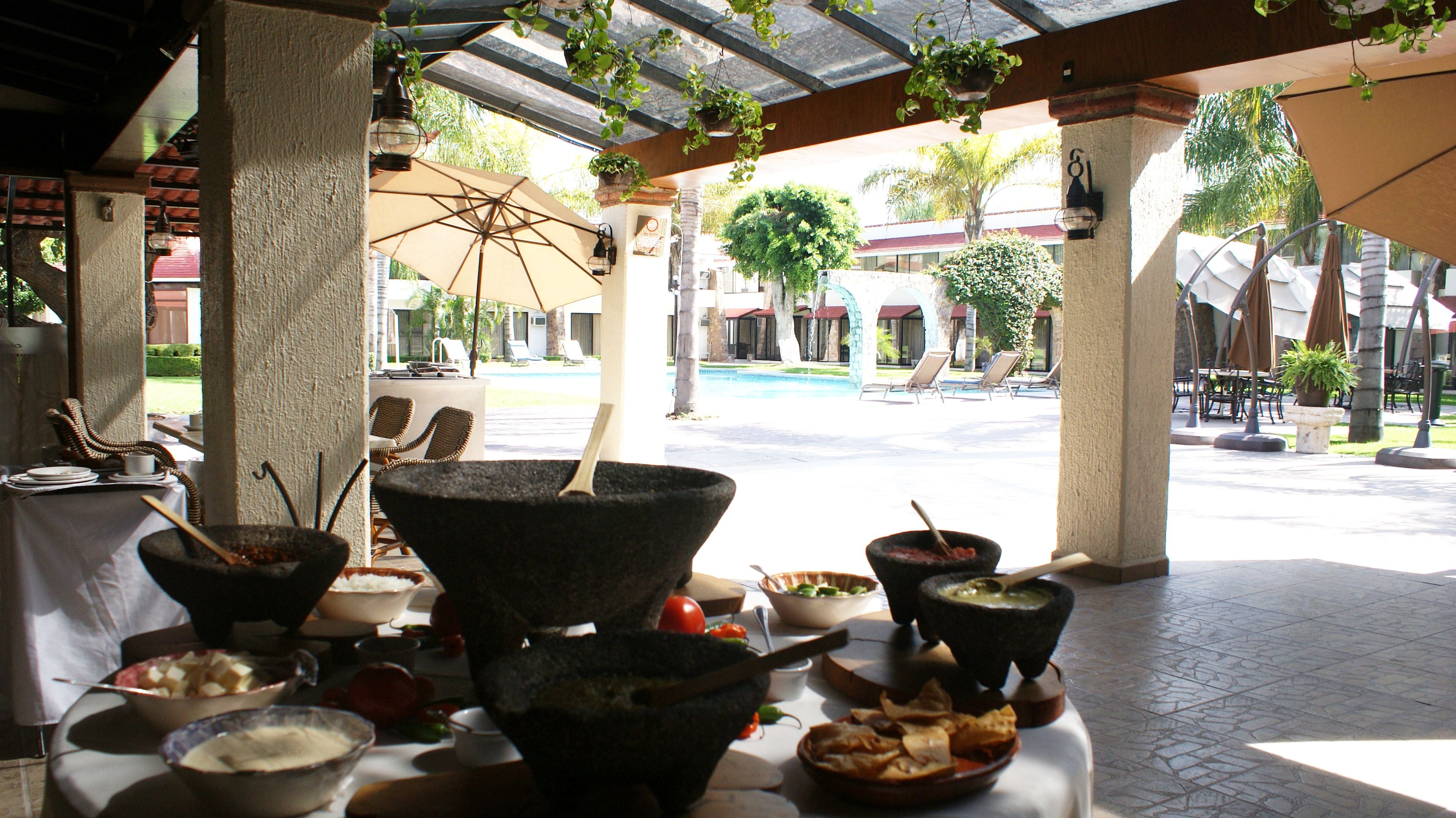 Zona de comedor al aire libre con cuencos y platos de piedra, rodeada de sombrillas y sillones, con vista a una piscina y edificios de un complejo turístico.