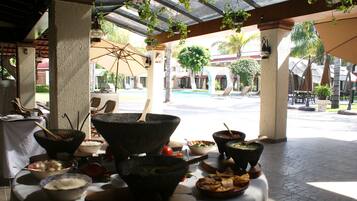 Zona de comedor al aire libre con cuencos y platos de piedra, rodeada de sombrillas y sillones, con vista a una piscina y edificios de un complejo turístico.