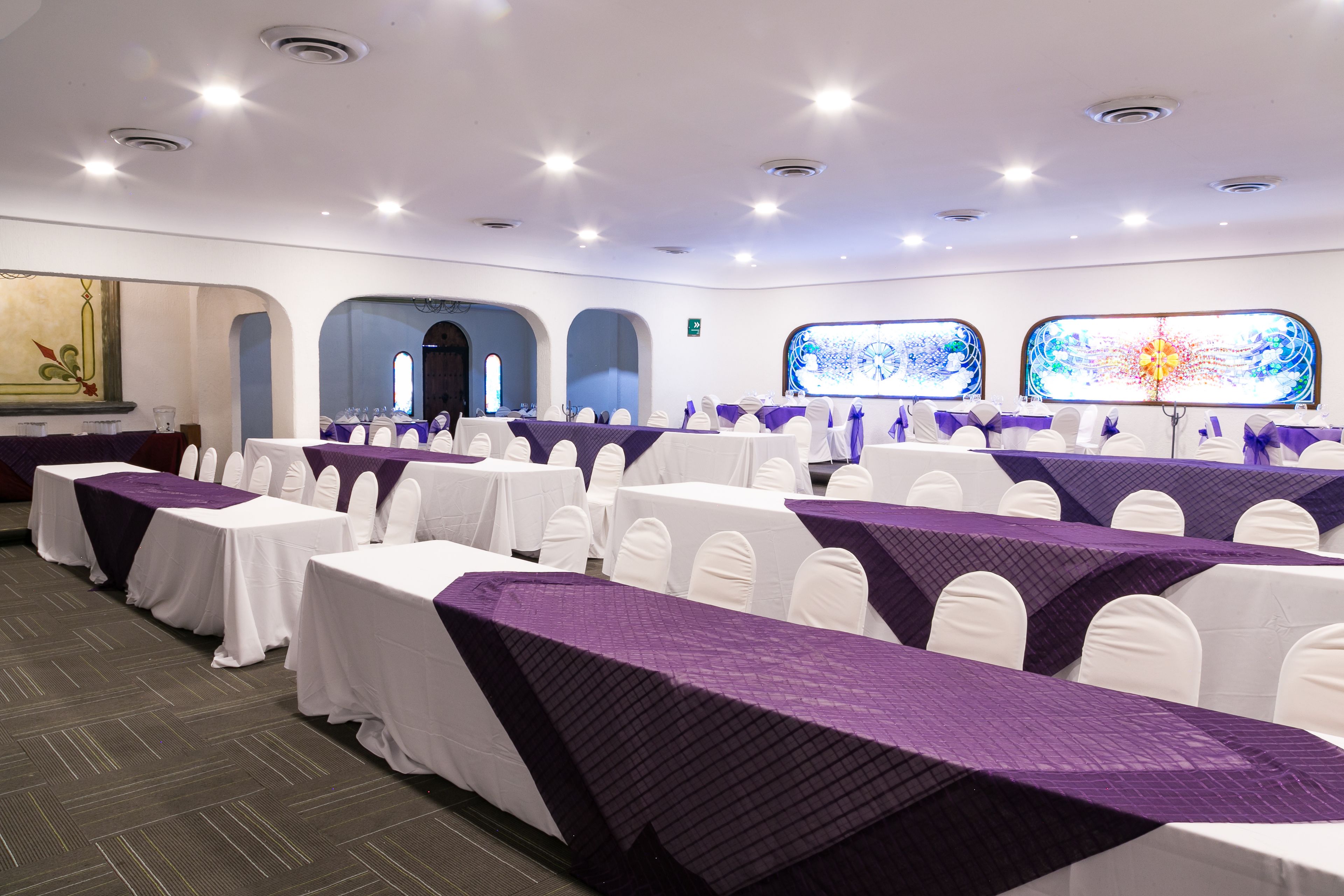 A conference room with rows of tables covered in white tablecloths and purple runners, white chairs, and large windows with decorative glass panels.