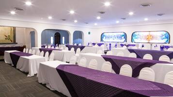 A conference room with rows of tables covered in white tablecloths and purple runners, white chairs, and large windows with decorative glass panels.
