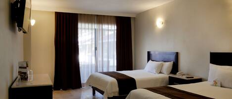A hotel room with two beds, a flat-screen TV mounted on the wall, a bedside table with a lamp, and a window with curtains.