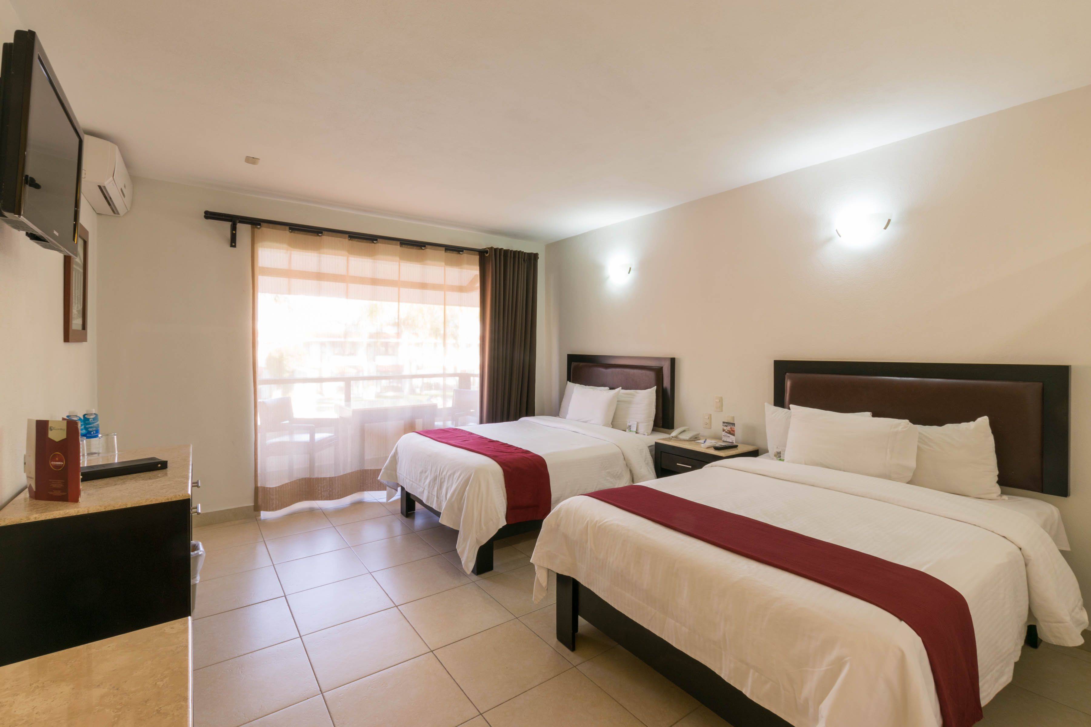 A hotel room with two beds, a desk, a TV, and a window with curtains.