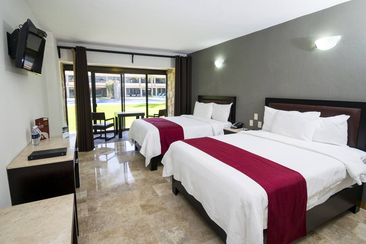 A hotel room with two beds, a desk, a TV, and a view of the outside.