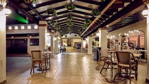 A spacious indoor area with wicker furniture and hanging plants, featuring a tiled floor and a high ceiling with exposed beams.