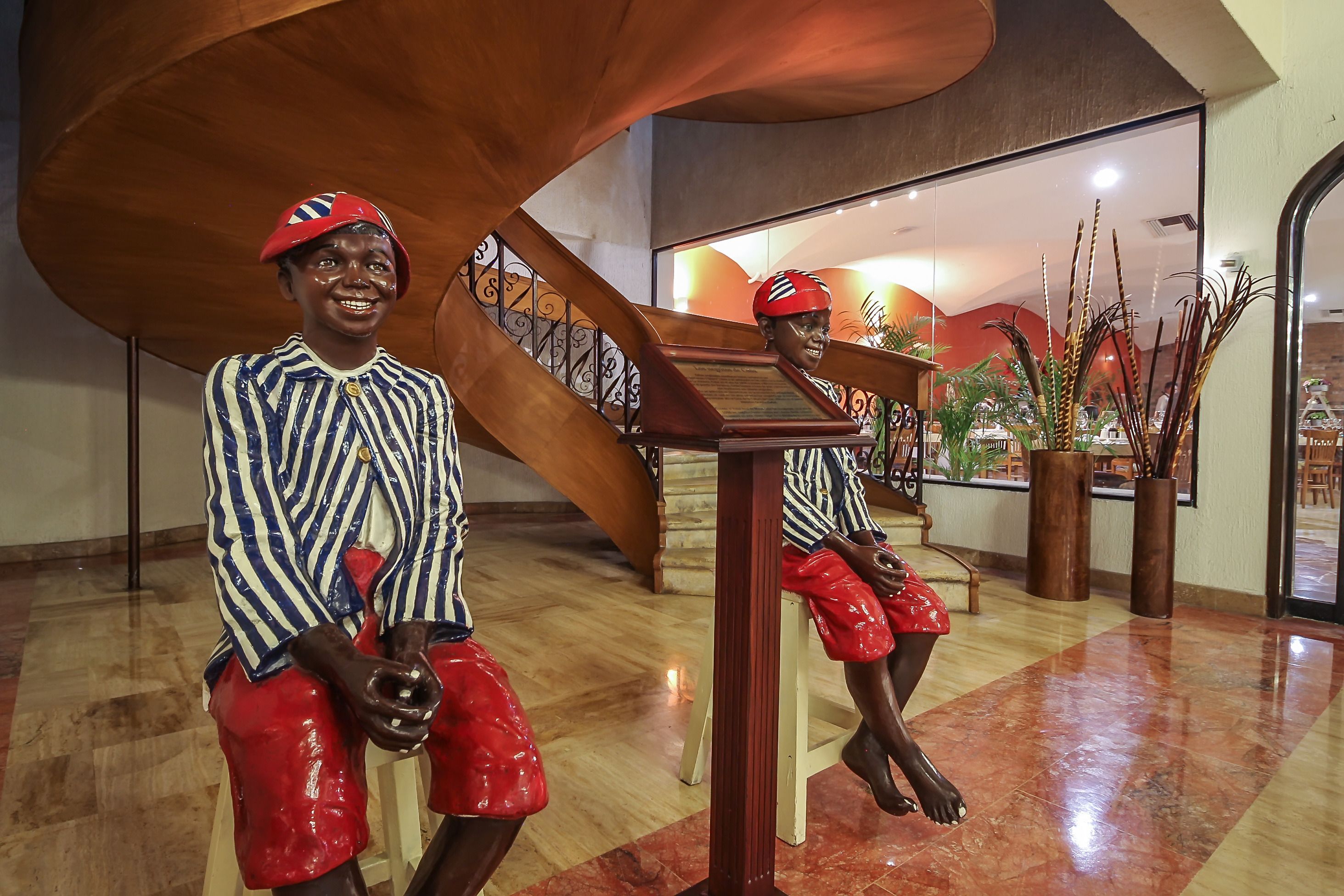 A sculpture of a person in a striped shirt and red shorts, seated on a wooden bench in a lobby with a polished floor and a staircase.