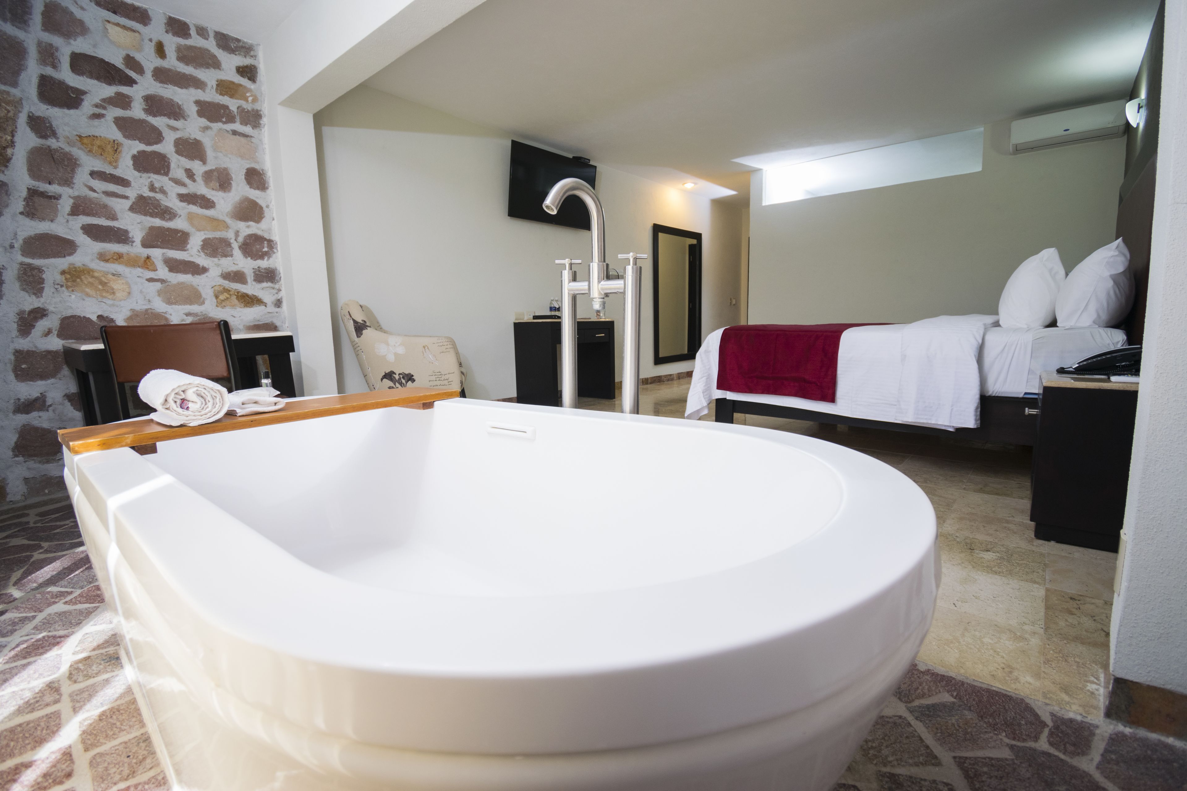A bathroom with a large bathtub, a stone wall, a bed with white bedding, and a television on the wall.