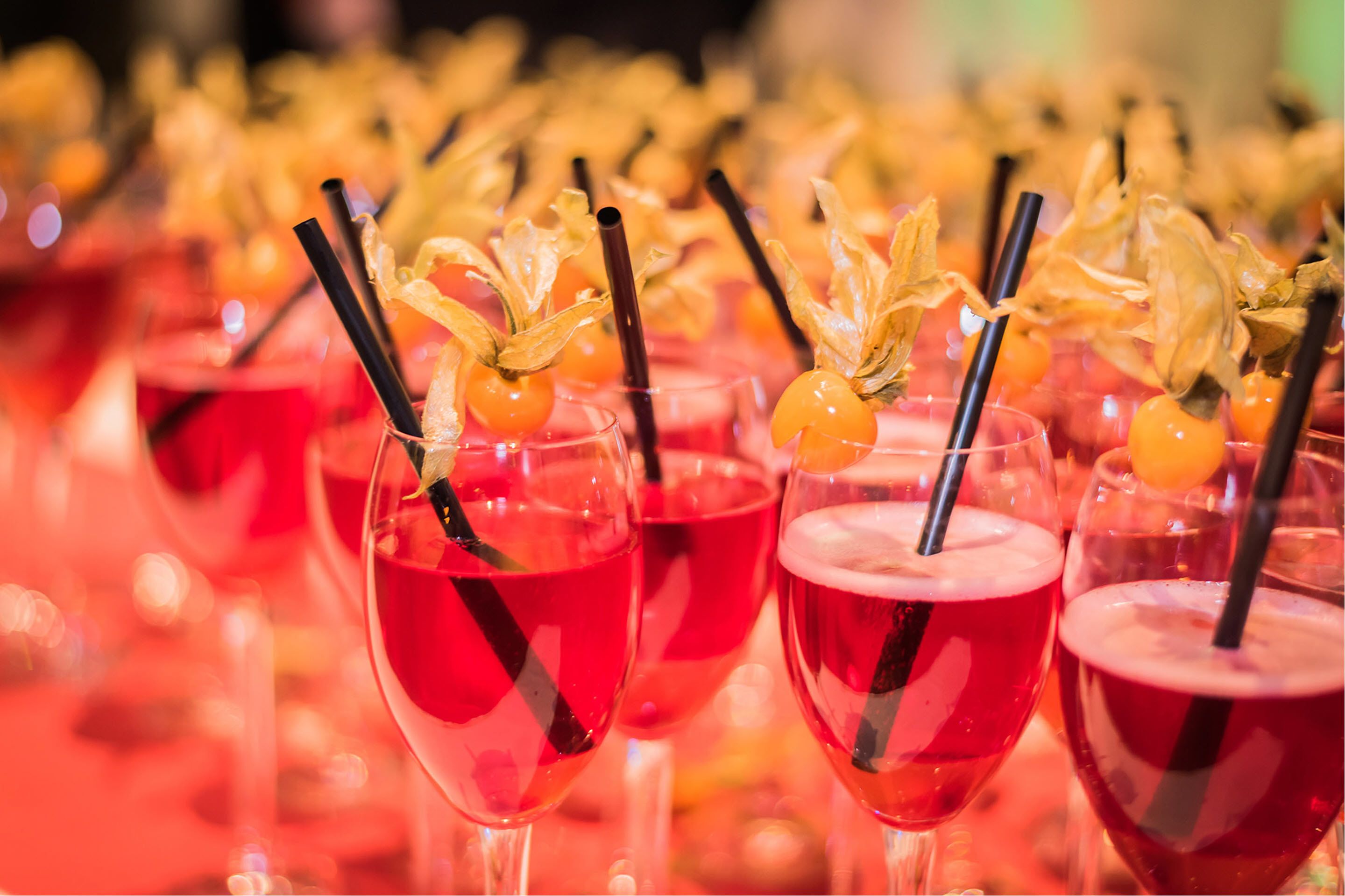 A row of red cocktails with black straws and decorative fruit garnishes.
