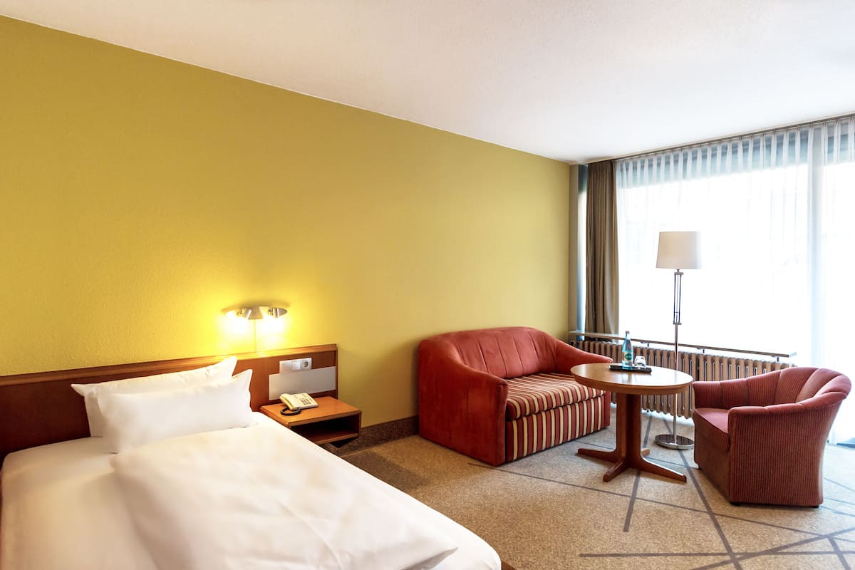 Una habitación de hotel con una cama, un sillón rojo, un sillón a rayas, una mesita y una lámpara.