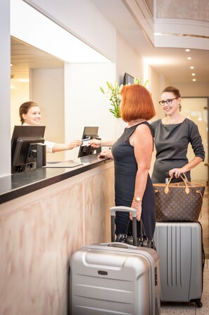 A reception desk with a woman and a man standing behind it, a laptop, and two suitcases. - Hotel Frankenland (Bad Kissingen)