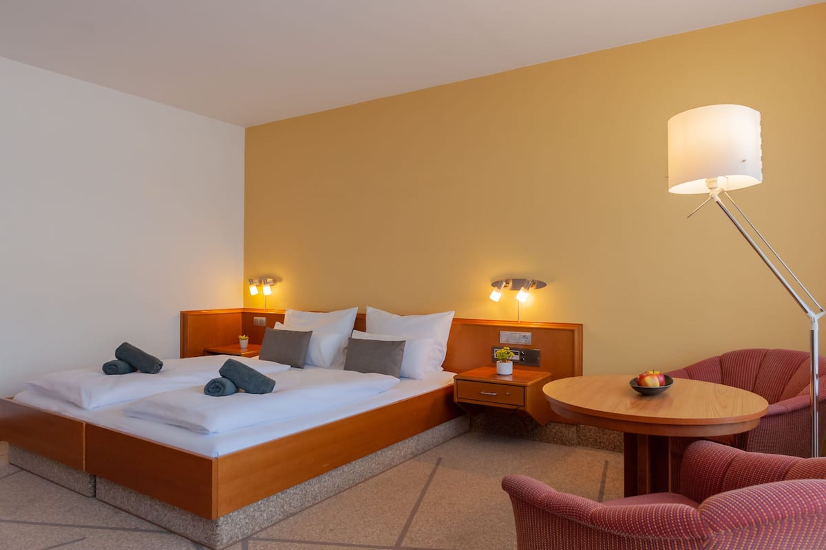 Una habitación de hotel con una cama doble, mesitas de noche con lámparas, una pequeña mesa redonda con un frutero y una silla tapizada de rojo.