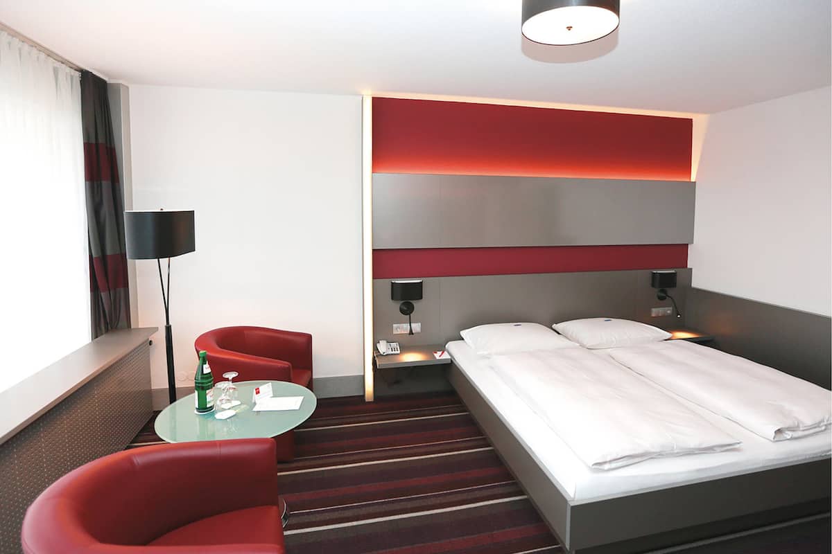Una habitación de hotel con una cama, un sillón rojo, una mesita con una botella y vasos, y una lámpara.