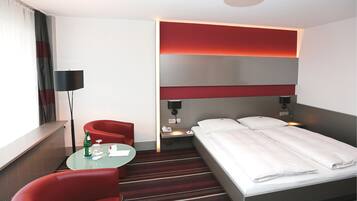 A hotel room with a bed, a red armchair, a small table with a bottle and glasses, and a lamp.