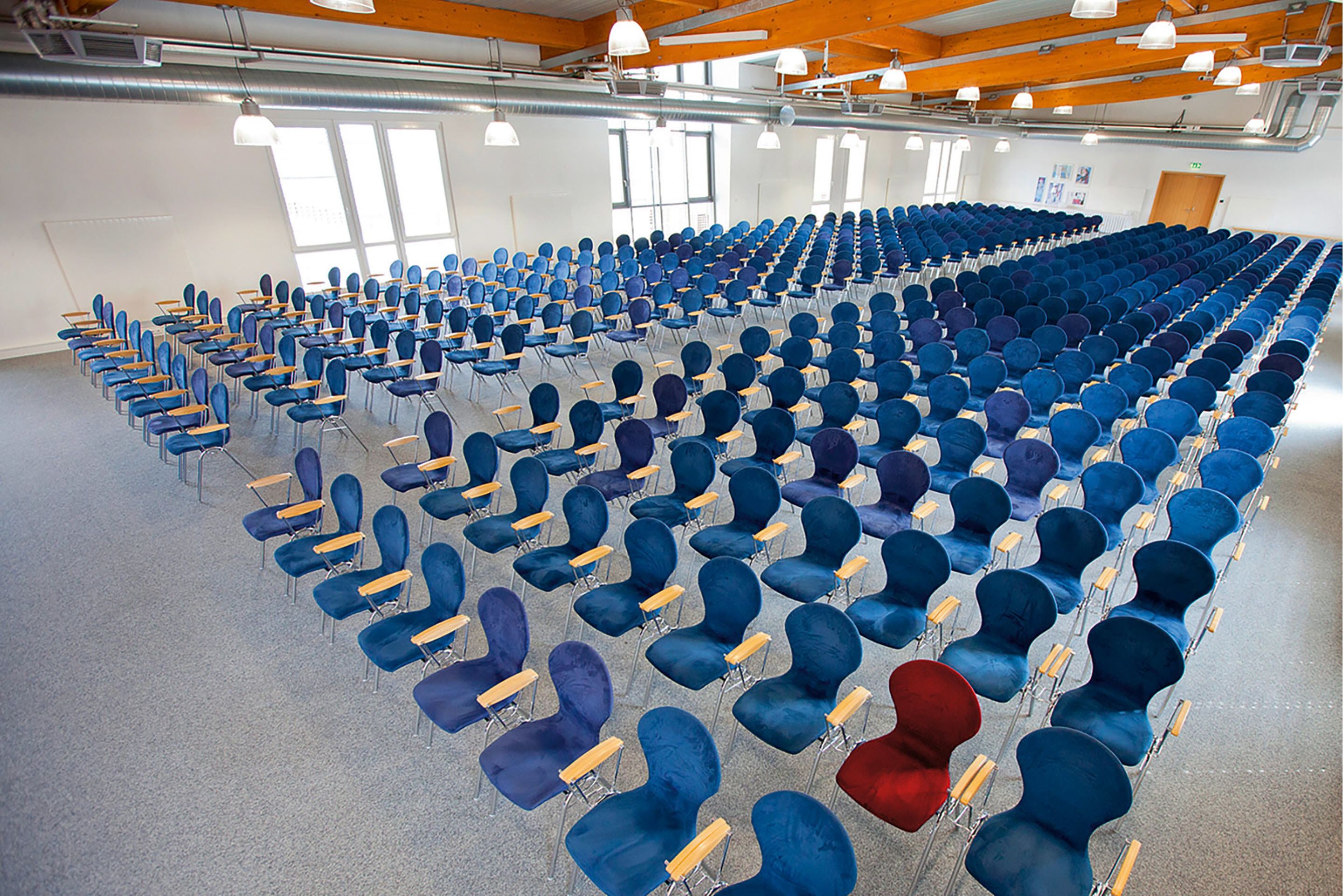 A large hall with rows of blue and red chairs, white pendant lights, and large windows.