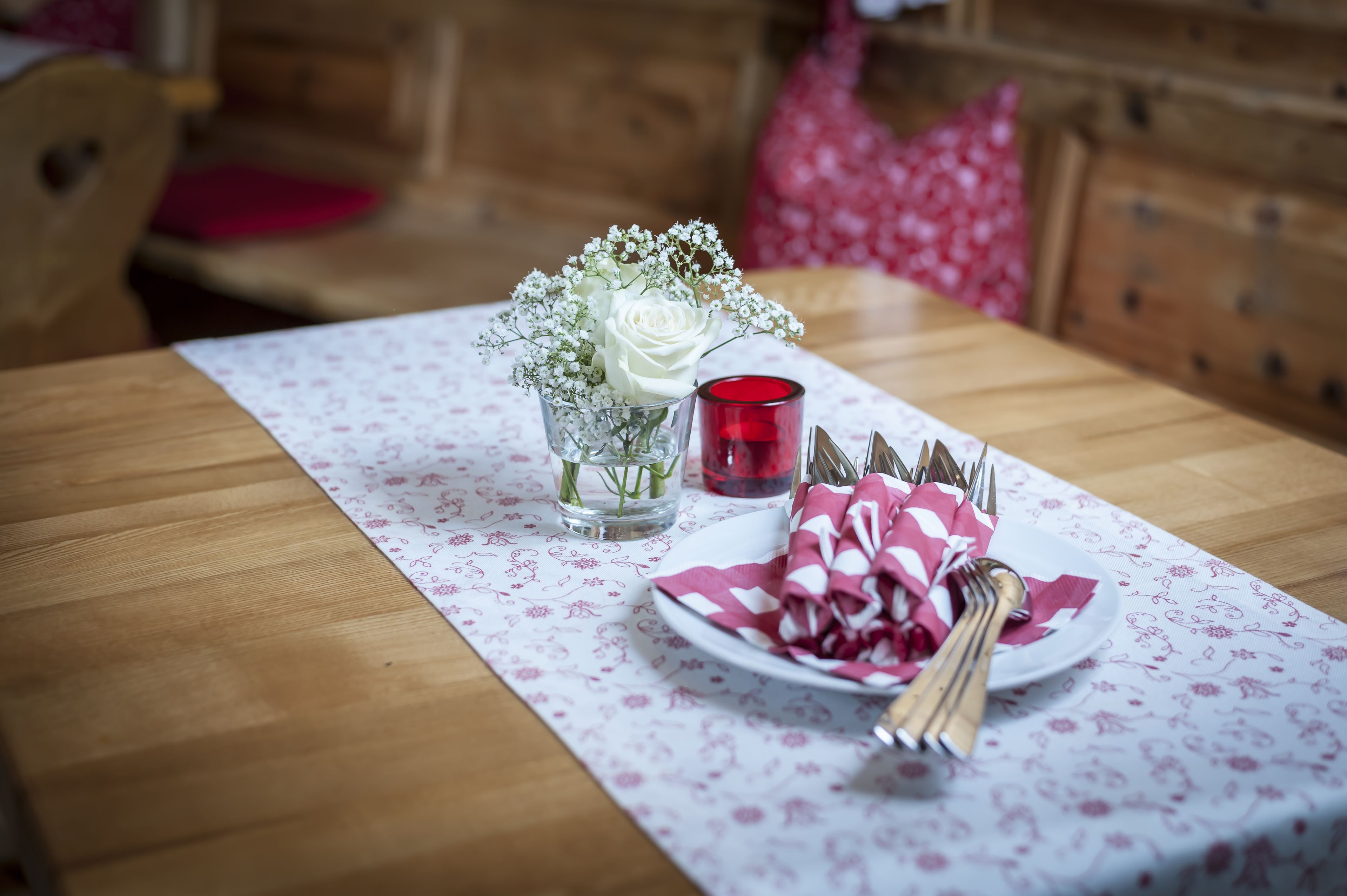 Una mesa de comedor con mantel floreado, un florero con flores blancas, una vela roja, y un plato con cubiertos y servilletas.