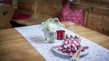 Una mesa de comedor con mantel floreado, un florero con flores blancas, una vela roja, y un plato con cubiertos y servilletas.