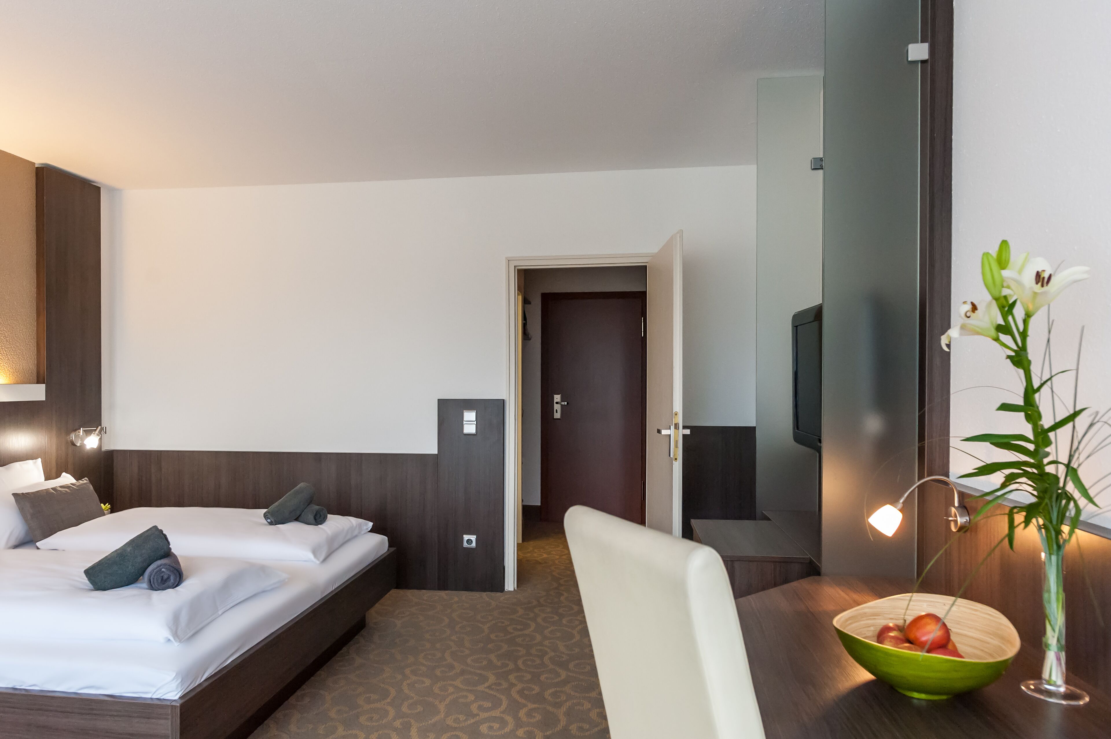 A hotel room with a bed, bedside table, lamp, and a bowl of fruit on a side table.