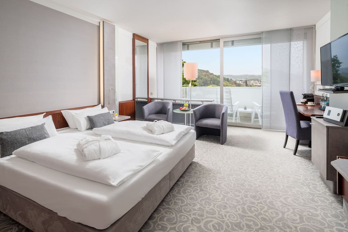 Habitación de hotel con una cama grande, dos sillones, un escritorio con televisor y un balcón con vistas.