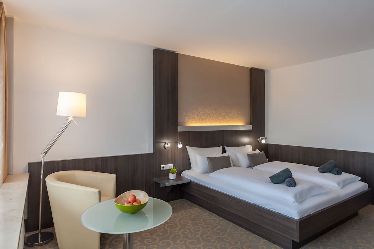 Habitación de hotel con cama, mesita de noche, lámpara, silla y una pequeña mesa con un cuenco.