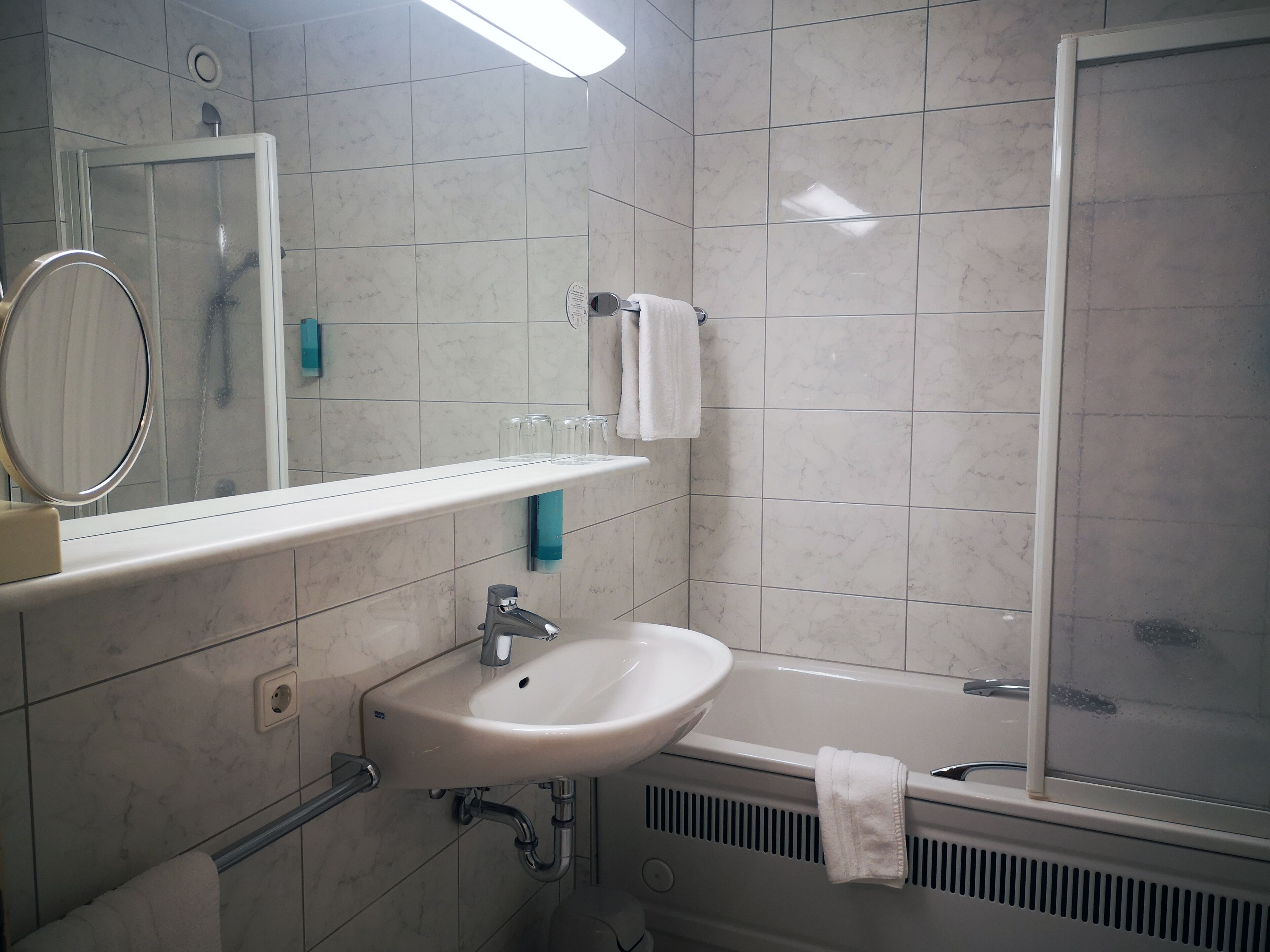 Une salle de bain avec un lavabo blanc, un miroir et une douche. On y trouve des bocaux en verre sur le comptoir et une serviette accrochée au mur.