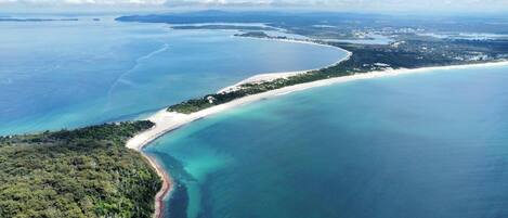 An aerial view of a coastal area with a sandy beach, clear blue waters, and dense greenery.
