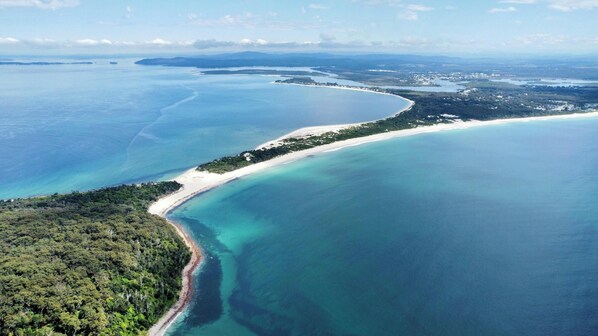 An aerial view of a coastal area with a sandy beach, clear blue waters, and dense greenery. - Anchorage Port Stephens (Corlette)