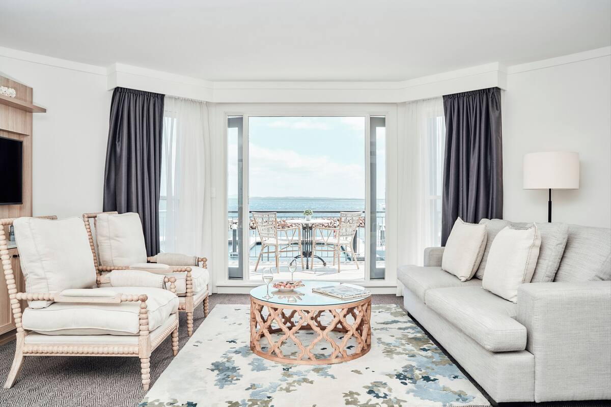A modern living room with a sofa, armchairs, and a coffee table. It features a large window with a view of the sea.
