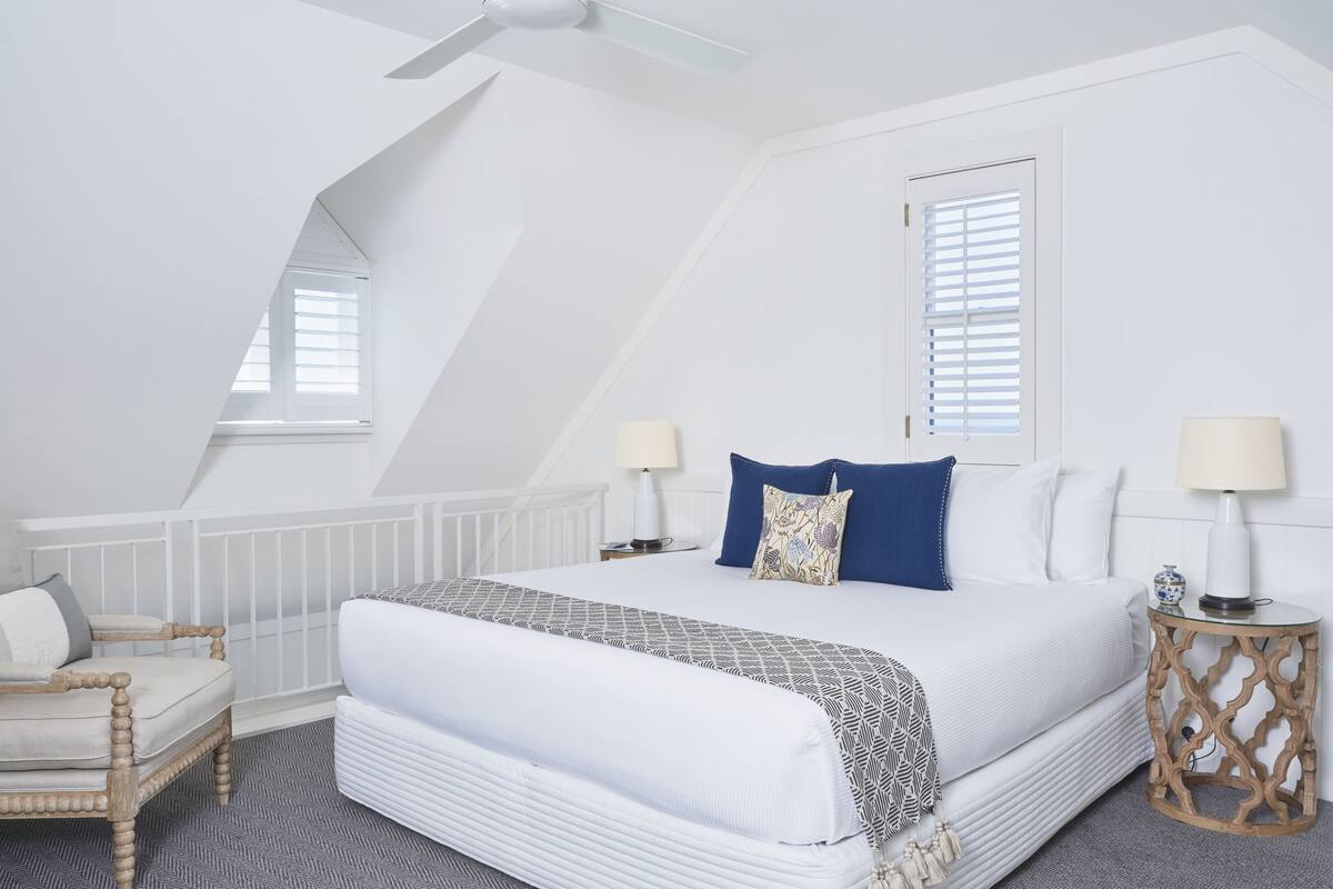 A bedroom with a large bed, two bedside tables, a chair, a window with blinds, and a ceiling fan.