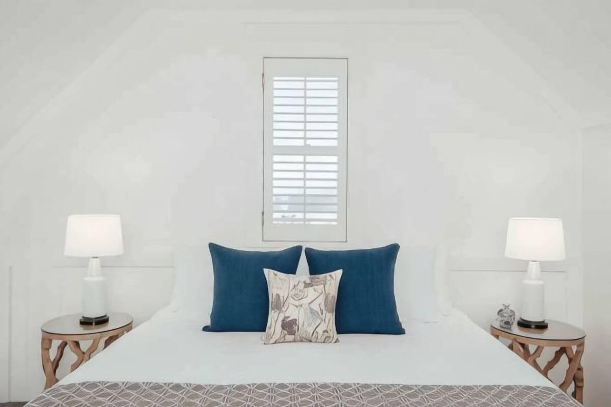 A bedroom with a bed, two bedside tables, a window with blinds, and a lamp on each table.