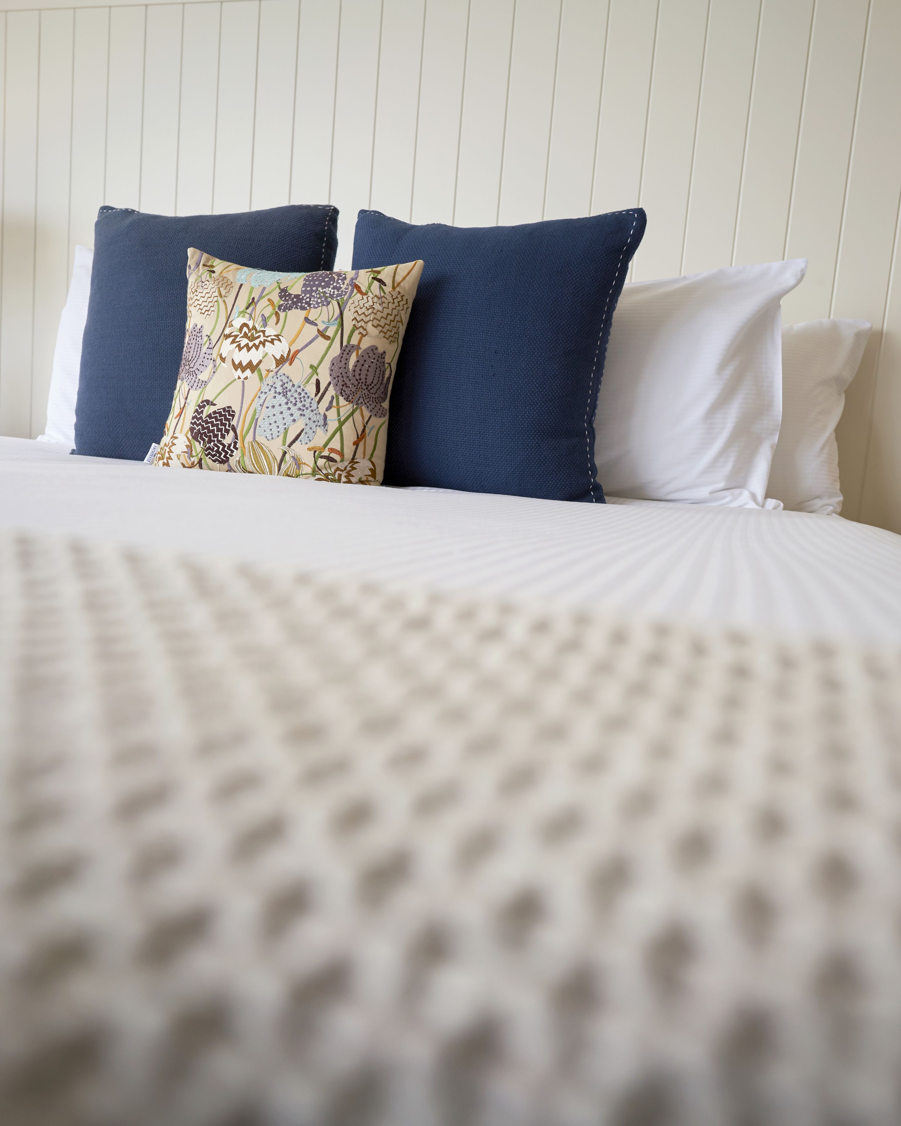 A neatly made bed with a patterned pillow, a solid blue pillow, and a plain white pillow.