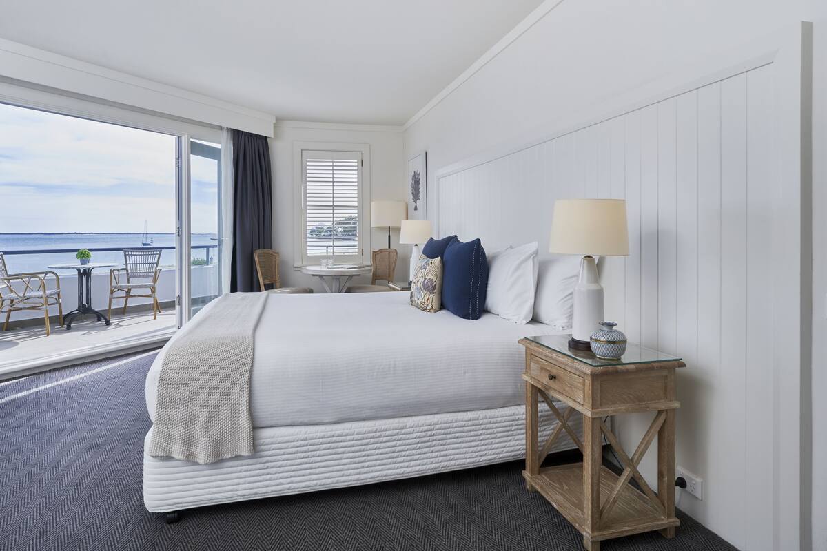 A hotel room with a large bed, a small table with chairs, a window with blinds, and a view of the sea.