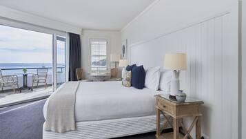 A hotel room with a large bed, a small table with chairs, a window with blinds, and a view of the sea.