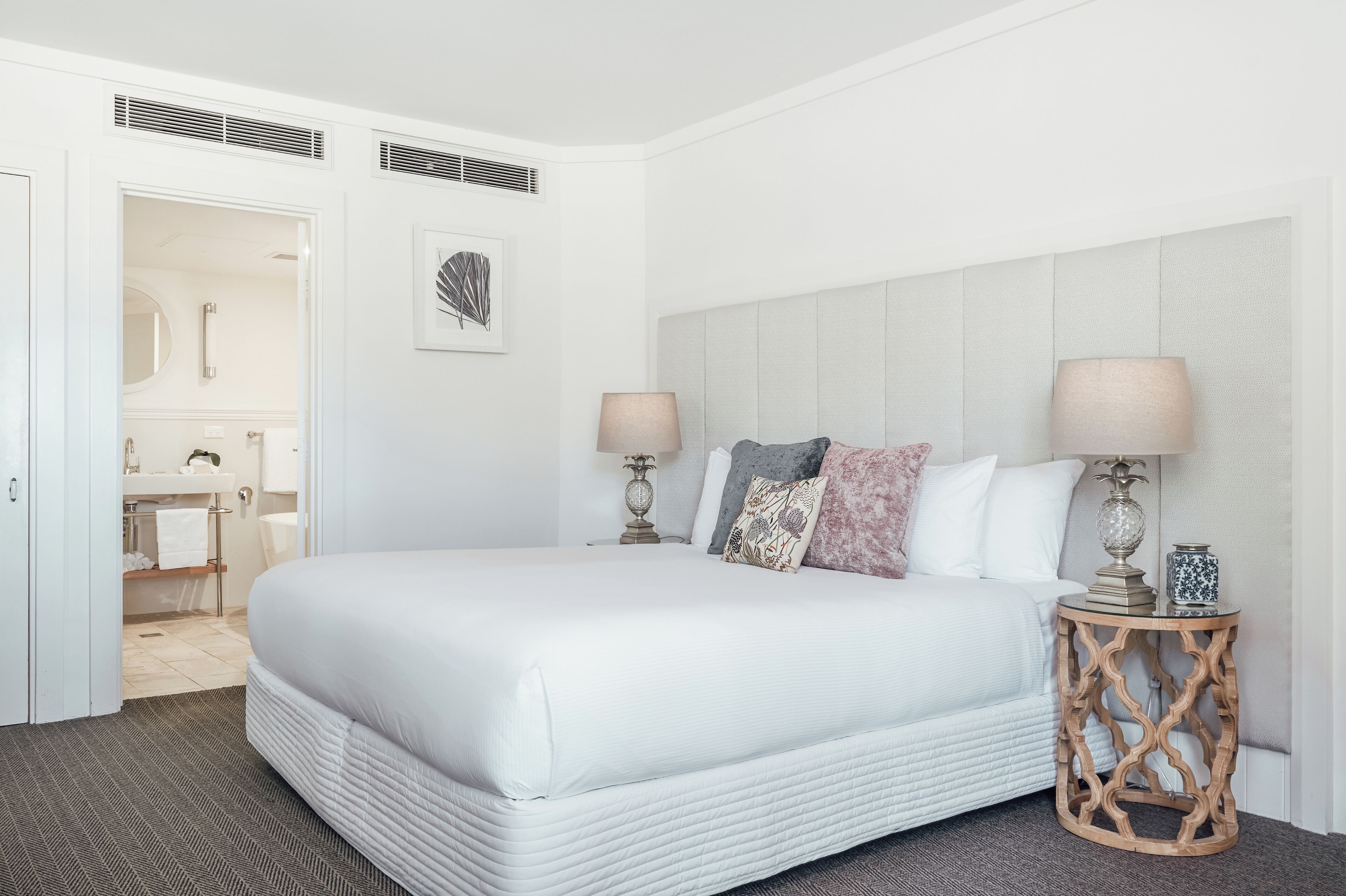 A hotel room with a large bed, two bedside lamps, a round wooden side table, and a view into the bathroom.