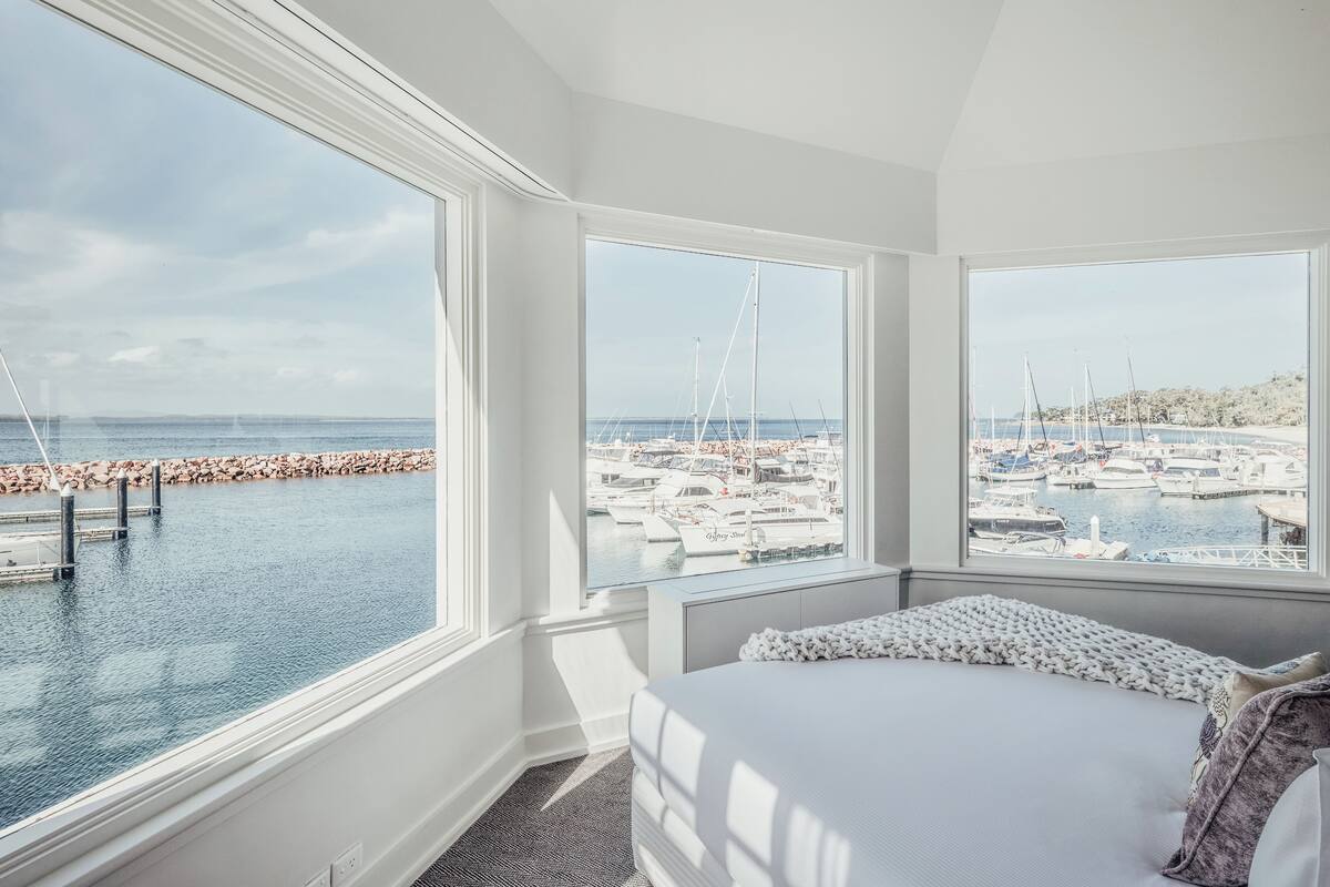 A bedroom with a large window overlooking a marina with boats and a rocky breakwater.